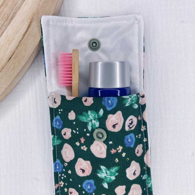 May include: A green fabric pouch with a floral print featuring pink and blue flowers. The pouch has a white lining and a snap closure. Inside the pouch are a pink and white toothbrush and a blue and silver bottle.