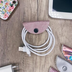 以下が含まれることがあります： スナップ留め具付きのピンクの革製のコードオーガナイザー。オーガナイザーは白い充電ケーブルを保持しています。