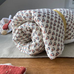 May include: A rolled up white and cream colored baby blanket with a small floral pattern in red and blue. The blanket has a gold ribbon sewn along the edge.
