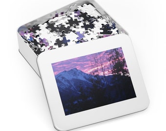Può includere: Una scatola di metallo con una scena di montagna sul coperchio. La scatola è aperta e piena di un puzzle.