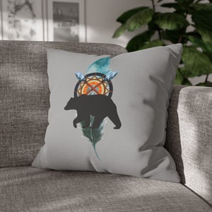 Puede incluir: Una almohada gris con un diseño de silueta de oso negro. El oso camina sobre una pluma azul y verde con un atrapasueños detrás.