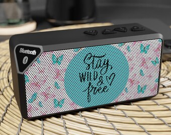 Puede incluir: Altavoz Bluetooth negro con un diseño floral y de mariposas. El altavoz tiene un círculo azul con el texto "Stay Wild & Free" en blanco.