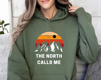 Può includere: Una felpa con cappuccio verde con una scena di montagna retrò e il testo "THE NORTH CALLS ME" stampato sul davanti.