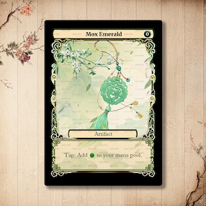 Puede incluir: Una carta verde y dorada de Mox Esmeralda de Magic: The Gathering con una ilustración de un colgante de jade verde. El texto de la carta dice "Mox Esmeralda 0 Artefacto Toca: Añade {G} a tu reserva de maná."