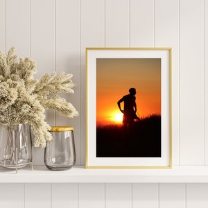 May include: A framed photograph of a person standing in a field at sunset. The person is silhouetted against the bright orange sky.