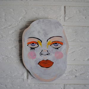 Op de afbeelding: Een handbeschilderde houten uitsparing in de vorm van een menselijk gezicht. Het gezicht is lichtgrijs geschilderd met feloranje oogschaduw, gele accenten, roze cirkelvormige blush op de wangen en opvallende oranje lippen. De ogen zijn zwart omlijnd.