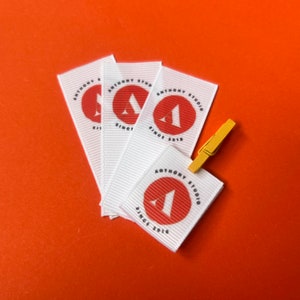 Könnte beinhalten: Vier weiße Stoff-Etiketten mit einem roten Logo und dem Text "Anthony Studio Since 2019".