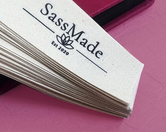 Op de afbeelding: Een stapel witte stoffen labels met zwarte tekst die "SassMade Est 2020" en een gestileerd bloemetjesontwerp zegt.