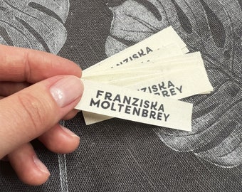 May include: Several off-white fabric labels with the text "FRANZISKA MOLTENBREY" printed in black. The labels are held in a hand, with a dark gray patterned background.