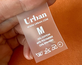 May include: A translucent clothing tag with the brand name "Urban" and website address. The tag displays the size "M" and fabric composition: 15% cotton, 60% polyamide, and 25% spandex. Washing instructions are also visible.