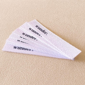 Könnte beinhalten: Eine Ansammlung weißer Stoffetiketten mit dem schwarzen Aufdruck "wonder.". Die Etiketten sind fächerförmig angeordnet und zeigen ihre gewebte Textur. Der Hintergrund ist hellbeige.
