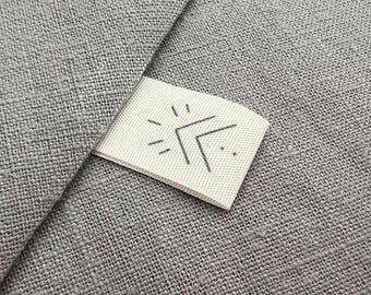 Pode incluir: Close-up de um tecido de linho cinza com uma etiqueta branca anexada. A etiqueta apresenta um design minimalista com um símbolo de sol e seta em cinza. O tecido tem uma aparência texturizada.