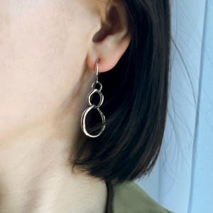 May include: Silver-toned drop earrings featuring a unique linked circle design. The earrings have a hook closure and a polished finish. The design consists of three connected circles, creating a modern and minimalist aesthetic. These earrings are a stylish accessory.