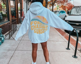 May include: A white hooded sweatshirt featuring a large circular graphic on the back. The design displays a stylized sunset in shades of orange, yellow, and blue, with the text "FOREVER CHASING SUNSETS" printed in bold yellow letters below the sun.