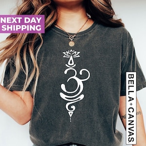 May include: A dark gray t-shirt with a white graphic of an Om symbol with a lotus flower above it. The text "BELLA + CANVAS" is on the right side of the shirt.