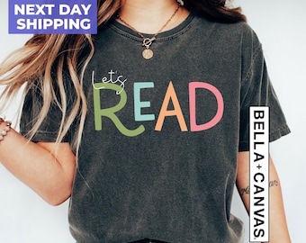May include: A dark gray t-shirt with the text "Let's Read" in colorful letters. The letters are in different colors, including green, blue, orange, and pink. The shirt is being worn by a person with long brown hair.