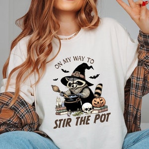May include: A white long-sleeved t-shirt with a graphic of a raccoon wearing a witch's hat and holding a broom. The text "On my way to stir the pot" is printed on the shirt.