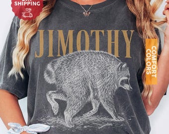May include: A charcoal gray t-shirt featuring the text JIMOTHY in large gold letters above a white line-art illustration of a raccoon walking. The shirt has a vintage-style washed appearance and short sleeves, showcasing a detailed animal graphic.