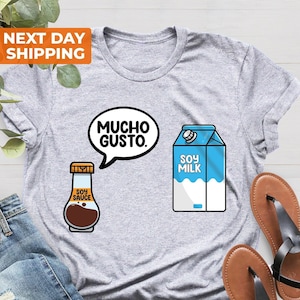 Peut inclure: Un t-shirt gris avec une bouteille de sauce soja et un carton de lait de soja. La bouteille de sauce soja porte l'inscription "Soy Sauce". Le carton de lait de soja porte l'inscription "Soy Milk". Le t-shirt porte également l'inscription "Mucho Gusto" dans une bulle de dialogue.