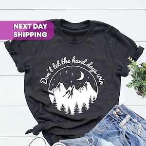 May include: Black t-shirt with a white graphic of mountains, a crescent moon, and stars. The text "Don't let the hard days win" is written around the graphic.