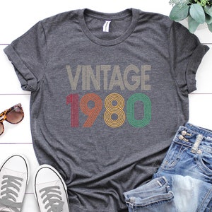 May include: A gray t-shirt with the text "Vintage 1980" in colorful retro-style lettering. The shirt is laid out on a white wooden surface with a pair of white sneakers, blue jeans, and sunglasses.