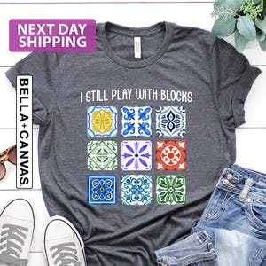 May include: Dark grey heather t-shirt with the text "I Still Play With Blocks" and a colorful tile design. The shirt is paired with blue jeans and white sneakers. The shirt is made of Bella+Canvas material.