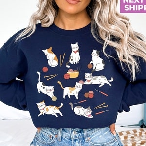 May include: A navy blue sweatshirt with a graphic of multiple cats playing with yarn and knitting needles. The cats are various colors and patterns.