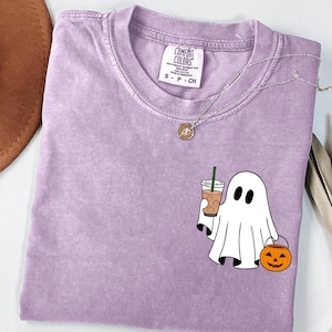 May include: A lavender Comfort Colors t-shirt featuring a white ghost graphic holding a coffee drink and a jack-o'-lantern. The shirt has a crew neck and short sleeves. A silver necklace with a pendant is also visible.