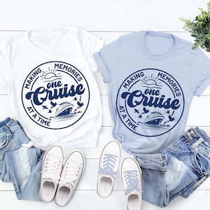 May include: Two t-shirts with a blue circle graphic that says "Making Memories One Cruise At A Time". One shirt is white and the other is light blue. Both shirts are laid out on a white wooden surface with a pair of white sneakers and two pairs of blue jeans.
