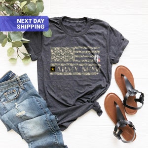 May include: A dark grey t-shirt with a camouflage American flag design and the text "Army Mom". A yellow star is above the text. A small tag with the word "David" is on the flag.