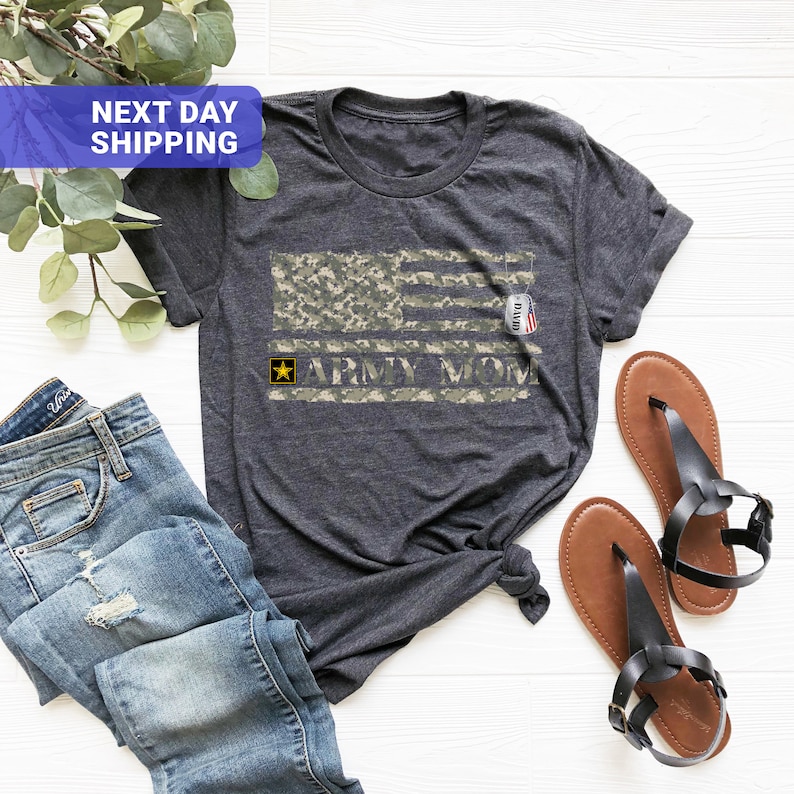 May include: A dark grey t-shirt with a camouflage American flag design and the text "Army Mom". A yellow star is above the text. A small tag with the word "David" is on the flag.