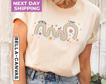 Może przedstawiać: Kremowy t-shirt z krótkim rękawem z grafiką przedstawiającą falistą, wielokolorową linię tęczy z małymi ilustracjami kwiatów oraz napisem "progress is not Linear" wydrukowanym ciemnoniebieskimi literami na klatce piersiowej.