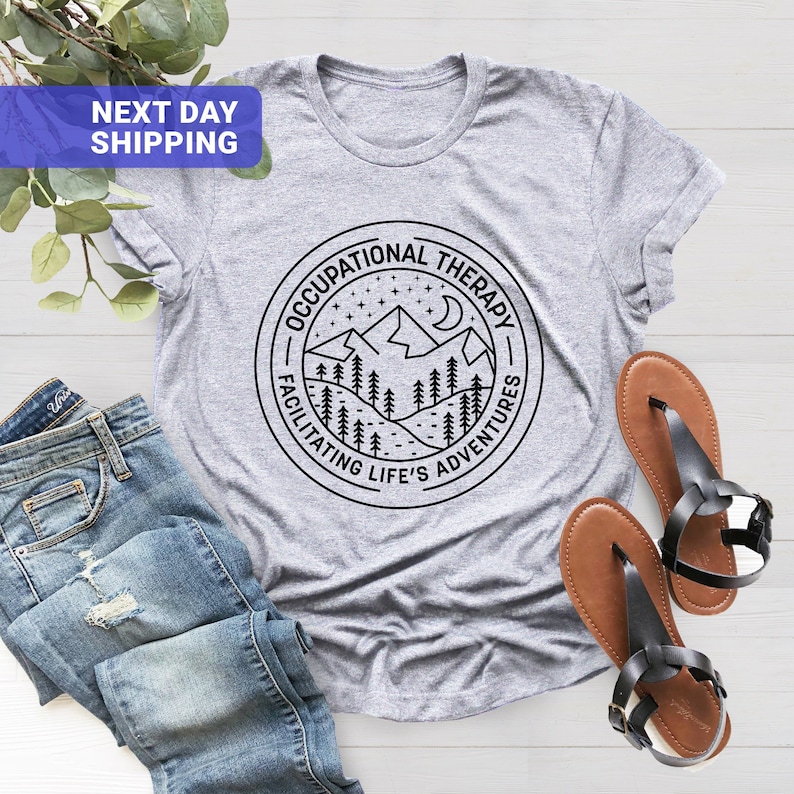 Op de afbeelding: Grijs T-shirt met een zwart-witte afbeelding van een bergketen met een halve maan en sterren. De afbeelding bevindt zich in een cirkel met de tekst "Occupational Therapy - Facilitating Life's Adventures".