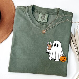 May include: Olive green t-shirt with a ghost graphic holding a coffee drink and a jack-o'-lantern bucket. The shirt has a Comfort Colors tag and a gold necklace. A brown hat and dried leaves are in the background.
