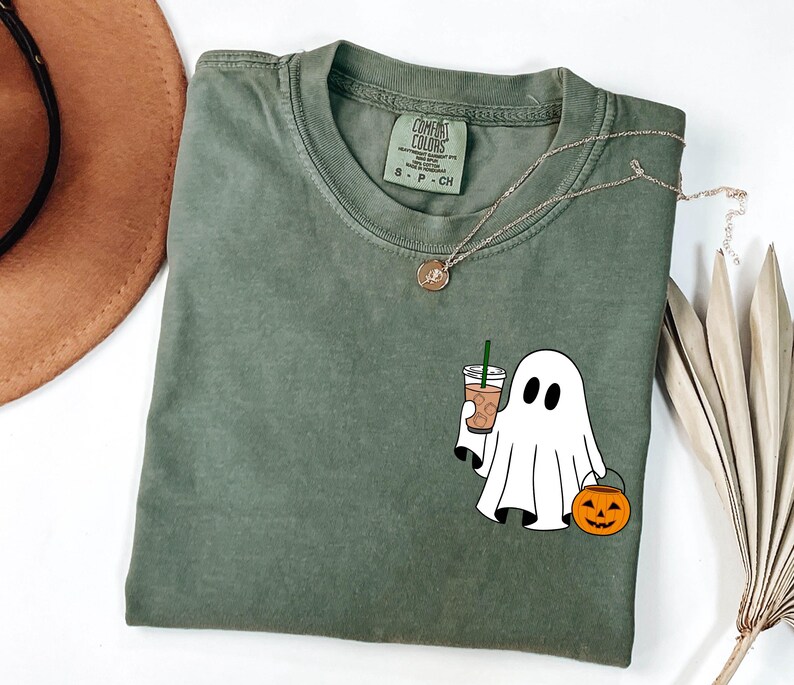 May include: Olive green t-shirt with a ghost graphic holding a coffee drink and a jack-o'-lantern bucket. The shirt has a Comfort Colors tag and a gold necklace. A brown hat and dried leaves are in the background.