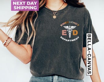 May include: A charcoal gray short-sleeve t-shirt featuring a white caduceus symbol centered between the letters E and D, with the text EMERGENCY DEPARTMENT arched above and below. The shirt has a crew neckline and a vintage-style wash.