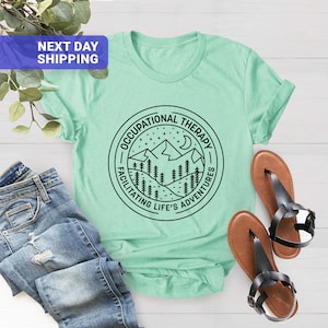 Op de afbeelding: Een lichtgroen T-shirt met een zwarte cirkelvormige afbeelding met een bergketen, sterren en een halve maan. De tekst "Occupational Therapy" staat bovenaan de cirkel en "Facilitating Life's Adventures" staat onderaan de cirkel.