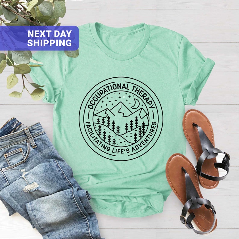 Op de afbeelding: Een lichtgroen T-shirt met een zwarte cirkelvormige afbeelding met een bergketen, sterren en een halve maan. De tekst "Occupational Therapy" staat bovenaan de cirkel en "Facilitating Life's Adventures" staat onderaan de cirkel.