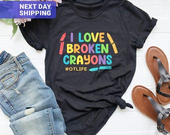 May include: A black short-sleeved t-shirt featuring the colorful text I LOVE BROKEN CRAYONS and the hashtag #OTLIFE. The design includes illustrations of broken crayons in red, yellow, and blue, set against a solid black fabric background.