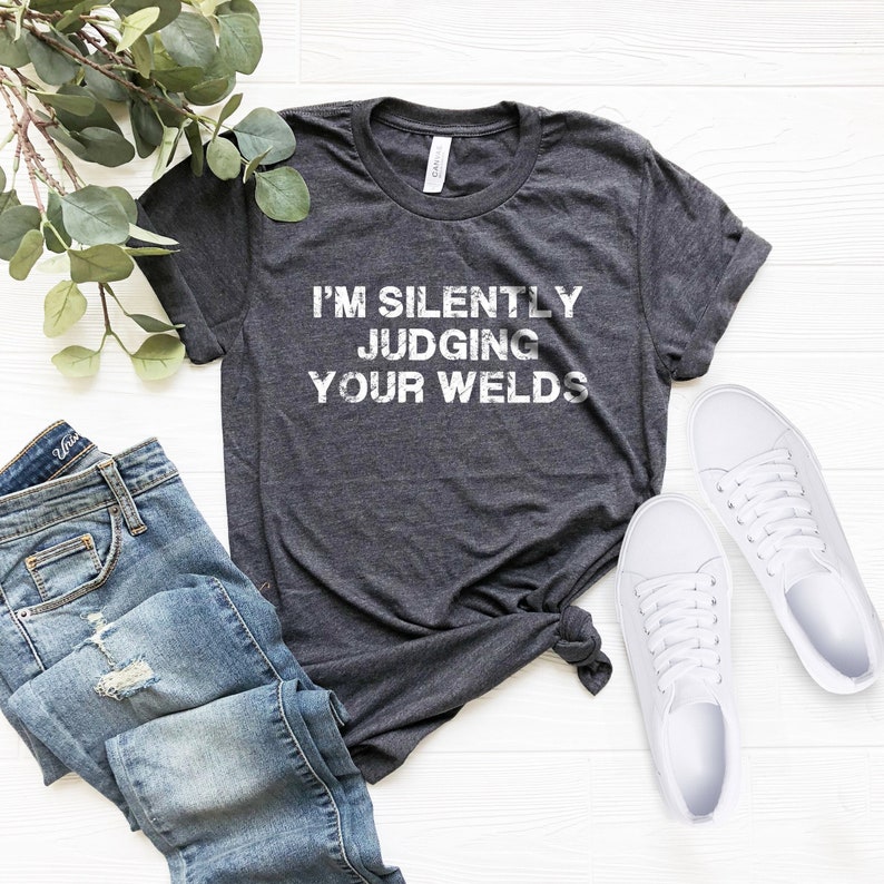 以下が含まれることがあります： 白い文字で「I'm silently judging your welds」と書かれた濃いグレーのTシャツ。Tシャツは折り畳まれて、白い木製の表面に置かれ、白いスニーカーと青いジーンズが置かれている。