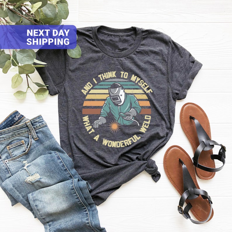 May include: A dark grey t-shirt with a retro-style graphic of a welder with the text "And I think to myself what a wonderful weld".