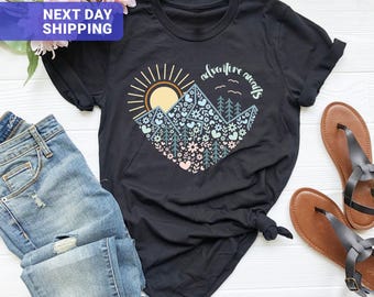 May include: A black short-sleeved t-shirt featuring a graphic of a heart-shaped mountain range with a sun, pine trees, flowers, and the text "adventure awaits" in a light blue, yellow, and pink colour palette on the front chest.
