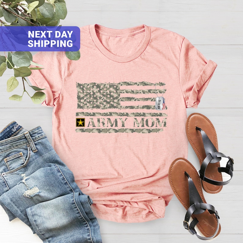 May include: A pink t-shirt featuring a camouflage American flag design and the text "Army Mom". A small silver tag with the name "David" is attached to the flag.