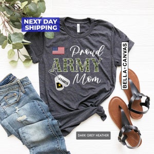 May include: Dark grey heather t-shirt with the text "Proud Army Mom" in white. The word "Army" is in camouflage print. A small American flag is in the top left corner. A camouflage dog tag with the name "James" is hanging from a chain.