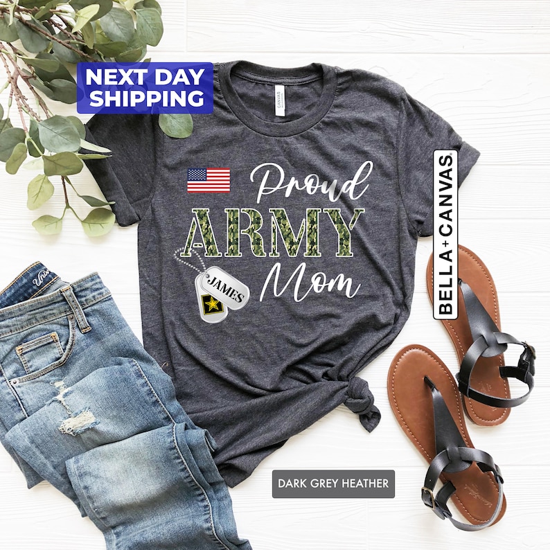 May include: Dark grey heather t-shirt with the text "Proud Army Mom" in white. The word "Army" is in camouflage print. A small American flag is in the top left corner. A camouflage dog tag with the name "James" is hanging from a chain.