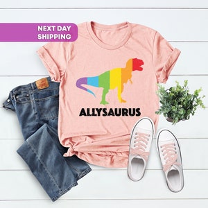 May include: Pink t-shirt with a rainbow dinosaur graphic and the text "ALLYSAURUS" printed on it. The shirt is laid out on a white wooden surface with a pair of white sneakers and blue jeans.