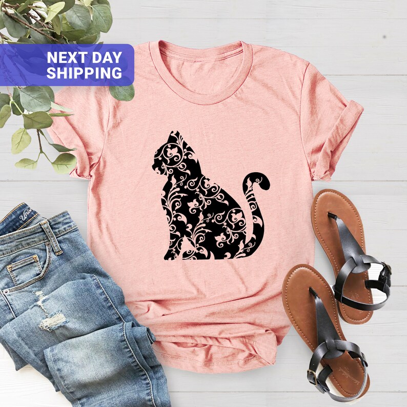 Puede incluir: Una camiseta rosa con una silueta negra de un gato con un patr&oacute;n floral. El gato est&aacute; sentado con la cola enroscada alrededor de su cuerpo.