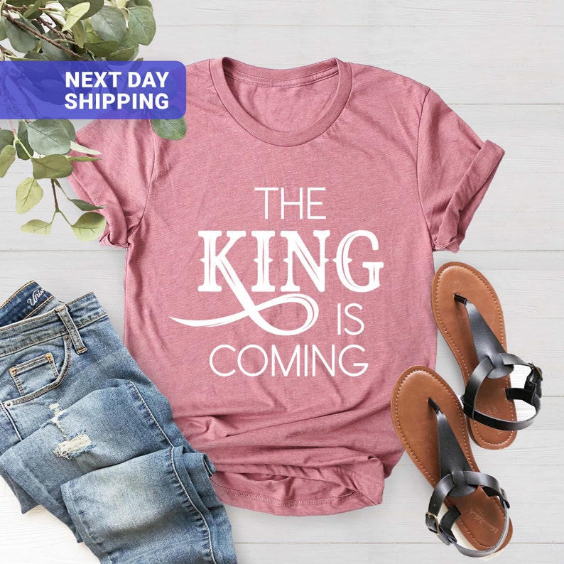 May include: A pink t-shirt with the text "The King is Coming" in white lettering.