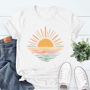 Op de afbeelding: Een wit T-shirt met korte mouwen en een grafische print van een gestileerde zon die boven het water uitkomt. Het ontwerp gebruikt een palet van gele, oranje en gedempte groene tinten. Het T-shirt ligt naast een blauwe spijkerbroek en witte sneakers.