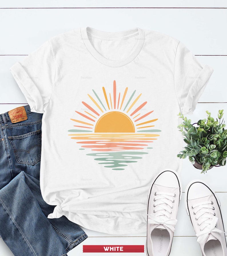 Op de afbeelding: Een wit T-shirt met korte mouwen en een grafische print van een gestileerde zon die boven het water uitkomt. Het ontwerp gebruikt een palet van gele, oranje en gedempte groene tinten. Het T-shirt ligt naast een blauwe spijkerbroek en witte sneakers.